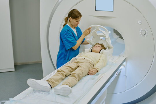 Caucasian female medical worker preparing Caucasian child for MRI scan, adjusting head support while child lying on examination table inside modern medical imaging machine - Powered by Adobe