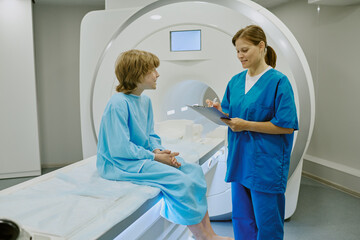 Caucasian boy sitting on MRI scanner bed talking with Caucasian female medical professional holding...