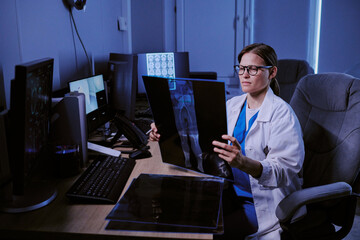 Caucasian young adult woman wearing glasses sitting at desk analyzing X-ray images in medical...