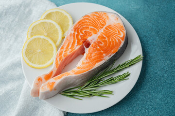 Closeup raw salmon steak with lemon slices and rosemary on white plate over blue background