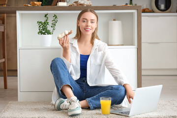 Young happy woman holding laptop, glass of juice and tasty toast with fresh butter in kitchen at...