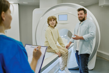 Caucasian middle aged man accompanying Caucasian child sitting on MRI scanner while female medical...