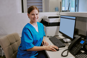 Caucasian young adult woman wearing medical scrubs sitting at desk working on computer in modern...