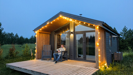 Person relaxes on porch of modern cozy glamping house at dusk. Glowing string lights creating warm,...