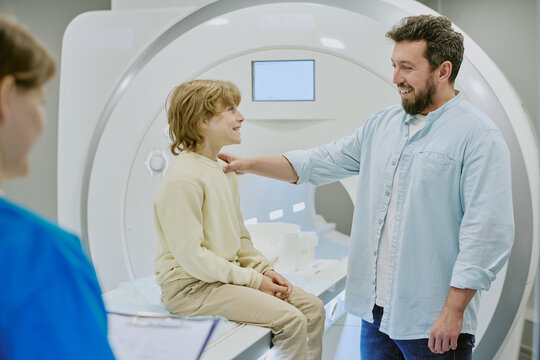 Caucasian child sitting on MRI scanner smiling at bearded Caucasian man standing beside, medical professional holding clipboard in foreground, hospital diagnostic setting
