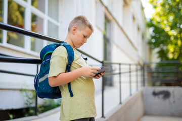 Focused schoolboy with backpack using digital tablet outside school building.
