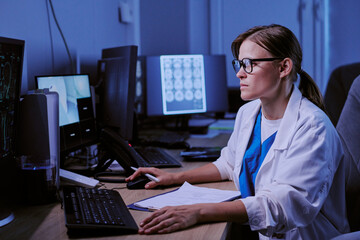 Naklejka na ściany i meble Caucasian young adult woman wearing glasses working at computer station in medical office analyzing brain scans on multiple monitors focusing on diagnostic imaging tasks
