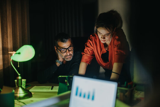A male and female coworker strategize together in a dimly-lit office setting, analyzing data from a laptop. Their focus and collaboration emphasize teamwork and late-night dedication to their business