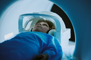 Caucasian child lying inside MRI scanner undergoing medical imaging procedure, wearing headphones and hospital gown, looking upward with calm expression during diagnostic test