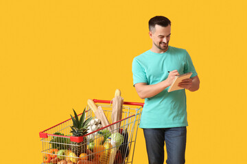 Young happy man with full shopping cart and notebook on yellow background