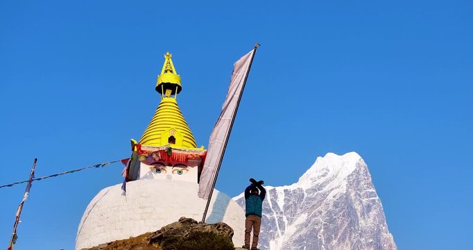 A male tourist waves to drone shot at a Buddhist stupa in Dingboche, Khumbu region, Nepal. Mt. Cholatse rises in backdrop with Buddhist flags, blue sky, and scenic views on the Everest trek serenity