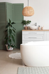 Interior of bathroom with bathtub, folding screen and chest of drawers near white wall