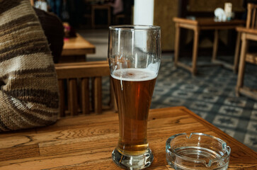 Amber ale beer fills pilsner glass, resting on wooden table with coaster and ashtray. Rustic pub features warm wooden furniture, knitted throw blanket, and patterned tiles, creating inviting