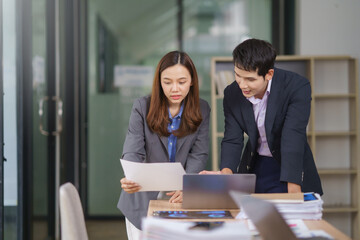 Asian businesspeople engaging in discussion over paperwork while collaborating on a laptop in a modern office environment filled with natural light