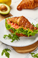 Delicious croissant sandwich with smoked salmon, avocado, and fresh greens being prepared on a wooden board