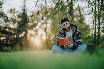 A man sits cross-legged on grass, engrossed in reading a book. The serene outdoor setting...
