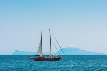 Naklejka premium A two-masted sailing yacht cruises the Tyrrhenian Sea in the Bay of Naples, Campania, Italy. The silhouette of the island of Capri is visible in the distance.