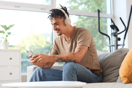 Young man with dreadlocks in headphones using mobile phone on sofa at home
