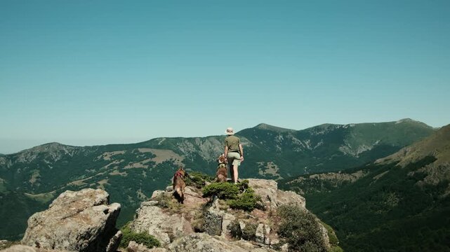 A woman with two dogs stands on the peak Babin Zub mountain in Stara Planina National Park, Serbia, enjoying the breathtaking view. Female tourist made arms out to the sides as embracing whole world