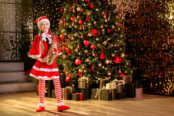 girl dressed as santa claus plays a saxophone near christmas tree