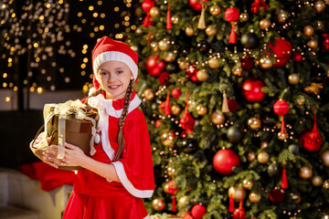 Girl in Santa costume holding Christmas gift box. Happy caucasian child with present near decorated tree. New year and winter holiday concept for greeting card