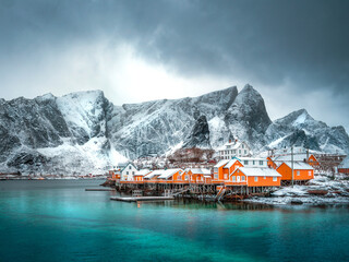 yellow huts of an Island with icy winter mountains in the background, foreground is clear blue water and there are dark storm clouds overhead. Winter in norway