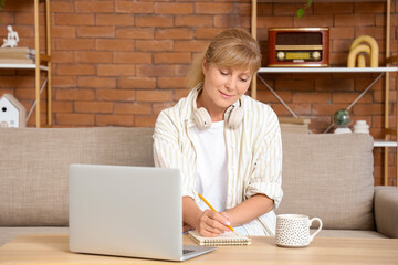 Mature woman with laptop writing in notebook on table at home