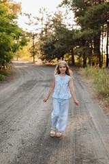 Naklejka premium Beautiful nine-year-old blonde girl with long hair in a blue suit, walking in nature in the forest. Summer, sunset