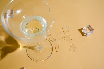 Broken glass with drink and ice cubes on sandy surface, sharp fragments scattered around