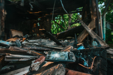 Ruins of burnt wooden house after fire, broken smartphone on the background. Burnt smartphone in ruins, symbol of fire hazards and carelessness