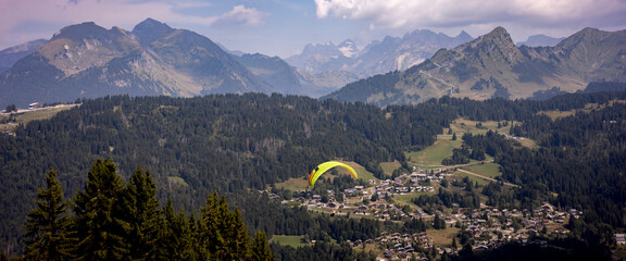Flying paraglider passing by in stunning French Alps scenery with Mont Blanc massive in the background and Les Gets town in valley below. Anamorphic cinematic panorama.