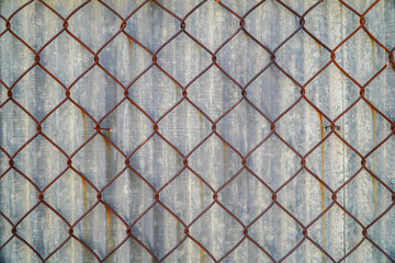 An aged chain-link fence with rust against a corrugated metal backdrop. The geometric pattern and...