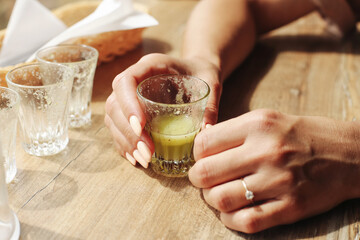 Woman hand holding a glass with green drink on wooden table. Empty glasses nearby with traces of previous drinks. Casual moment at outdoor cafe. Relaxing atmosphere with alcohol or herbal shot.