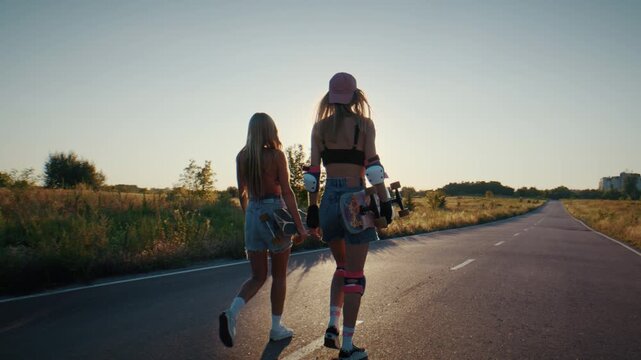 Sporty women walk on country road at sunset carrying surfskates