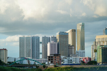 Fototapeta premium A cityscape featuring various modern buildings with diverse architectural styles set against a cloudy sky, with some buildings under construction near a bridge and greenery; a perfect stock image.