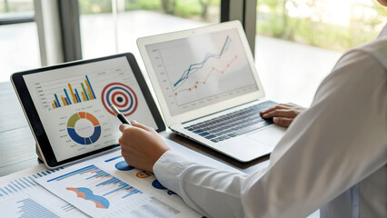 Professional analyzes financial data on laptop and tablet, charts and graphs for strategic planning in a modern, bright office space for success