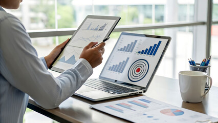 Professional analyzes financial data on laptop and tablet, charts and graphs for strategic planning in a modern, bright office space for success