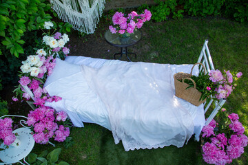 Bed with white bedding decorated with blooming pink and white peonies in a green garden