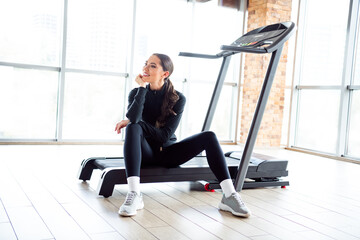 Athletic woman sitting near treadmill in stylish gym wearing activewear with bright daylight from...