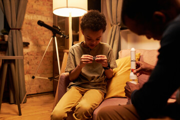 Modern African American man and his kid sitting together on couch in living room building toy space...