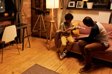 High angle of modern African American man and his son spending time together sitting on couch in...