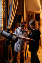 Vertical portrait of Caucasian woman and her daughter learning astronomy reading book about planets and stars and using telescope at home