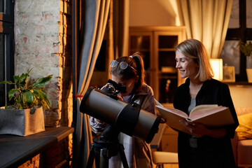 Horizontal medium of joyful Caucasian woman and her daughter learning astronomy reading book about planets and stars and using telescope at home