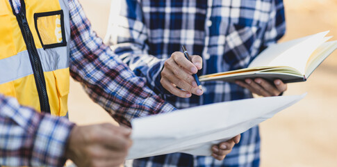 Two engineers are discussing and analyzing blueprints while taking notes on a construction site.