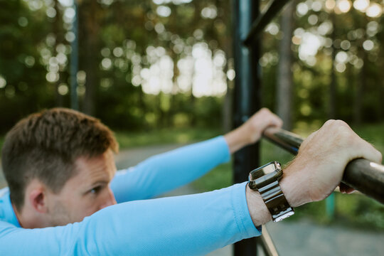 Caucasian young adult man gripping pull up bar outdoors, focusing on exercise with smartwatch on wrist, forest background visible, arms extended, preparing for workout