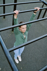 Caucasian young adult woman hanging from outdoor monkey bars, gripping metal bars with both hands,...