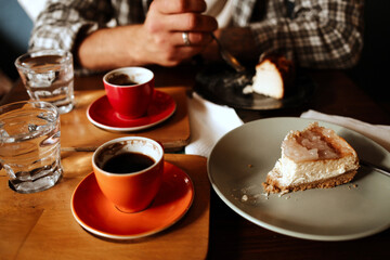 Two cups of espresso and a slice of cheesecake on a table with a man holding a spoon in the background. Cozy date in cafe scene