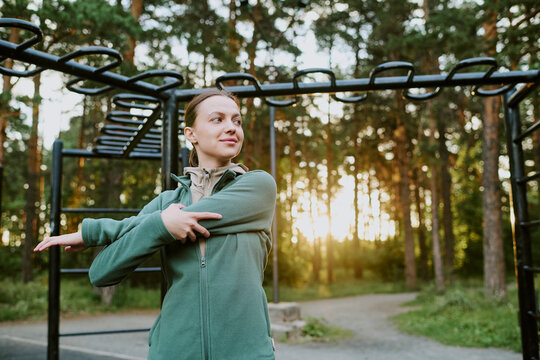 Caucasian young adult woman stretching arms outdoors near monkey bars in park, preparing for workout, looking away from camera with focused expression, surrounded by tall trees