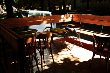 Outdoor cafe terrace with wooden tables, chairs, and cushions in sunlight under trees. Belgrade, Serbia