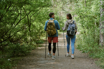 Young adult Caucasian man and young adult Caucasian woman hiking together on forest trail, carrying...